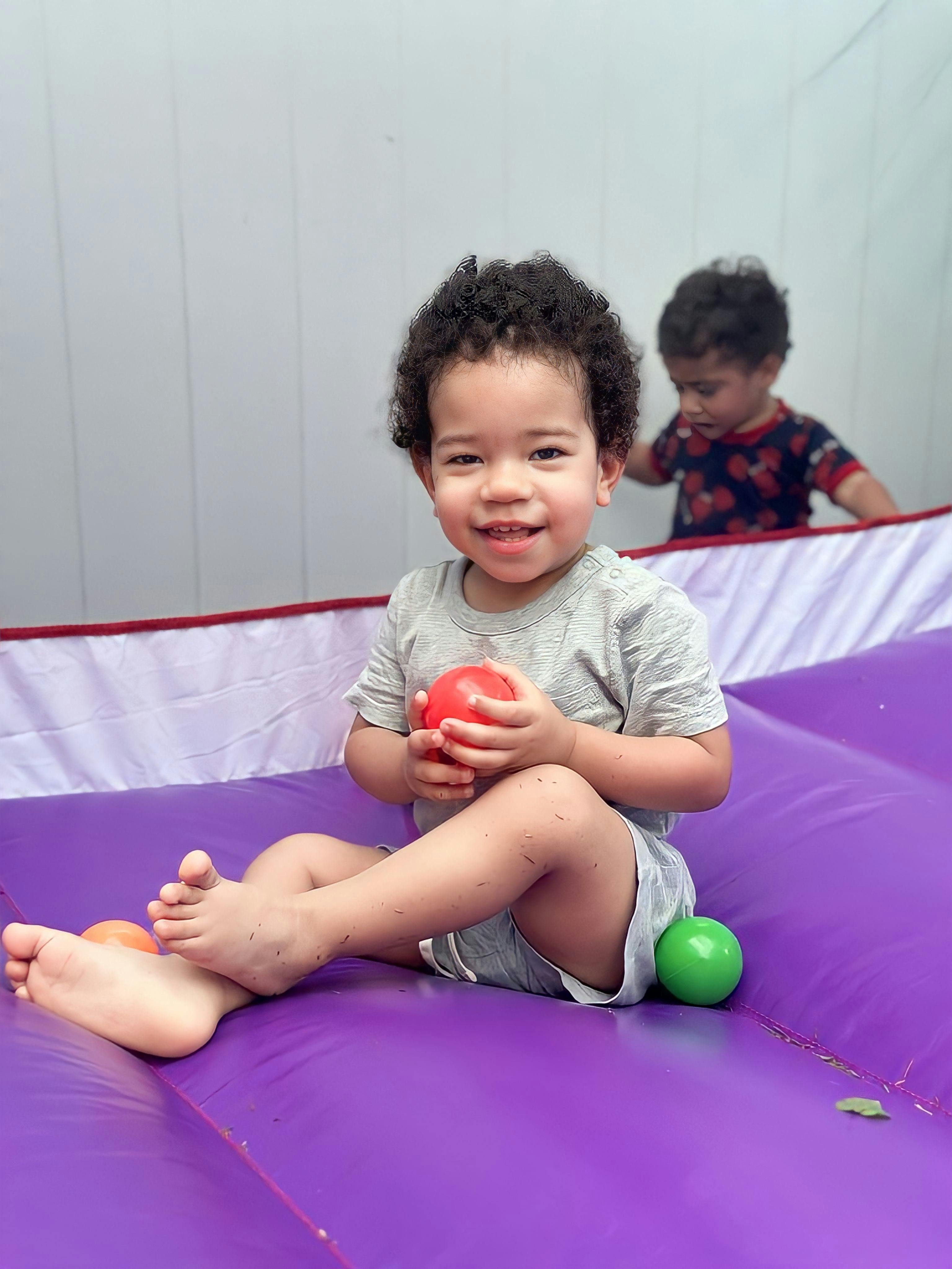Child sitting on purple surface holding a red ball, another child in background