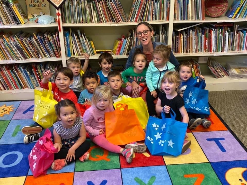 Adult and children sitting on a rug with colorful bags in front of bookshelves