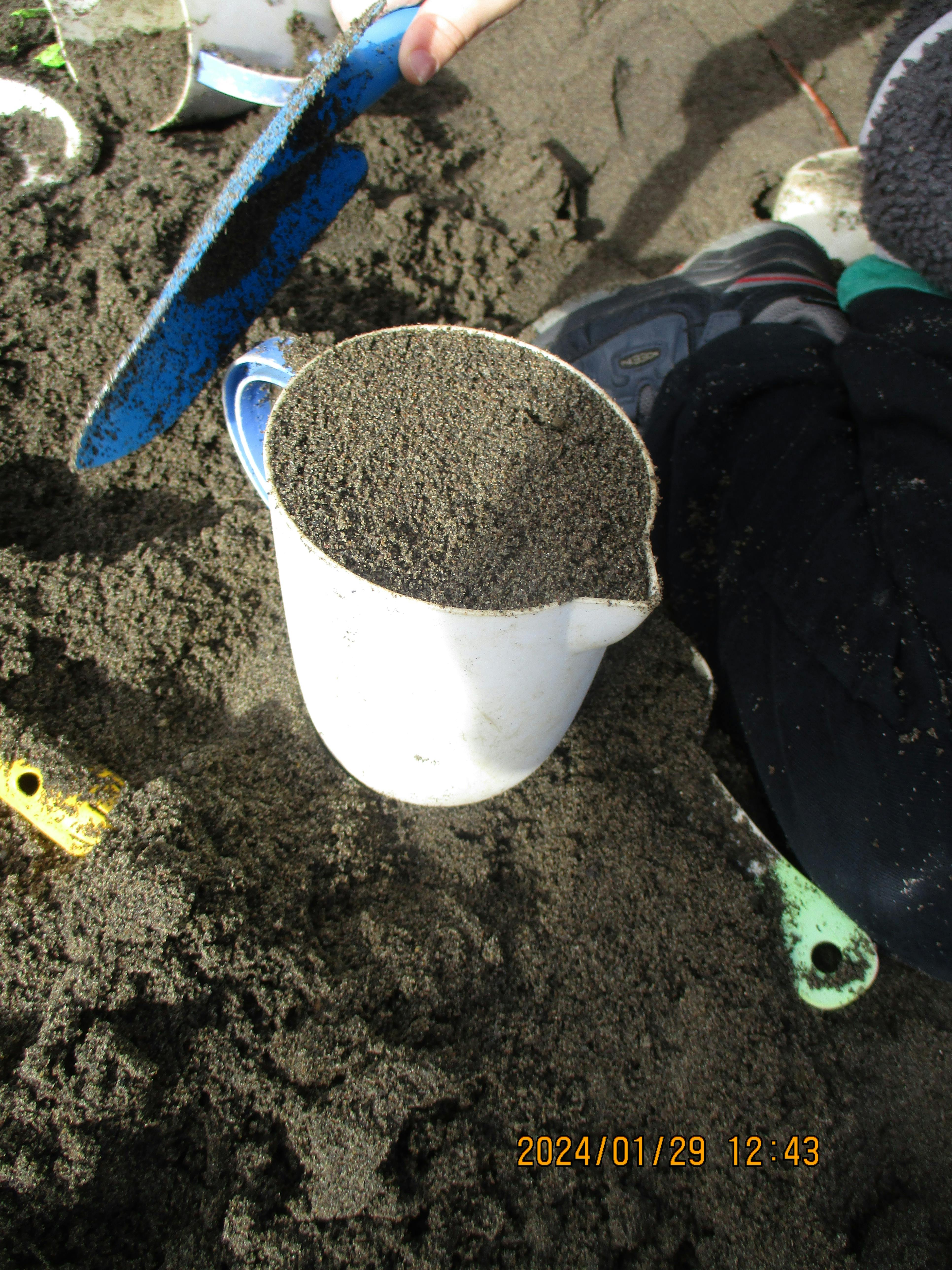 Person filling a white cup with sand using a blue shovel