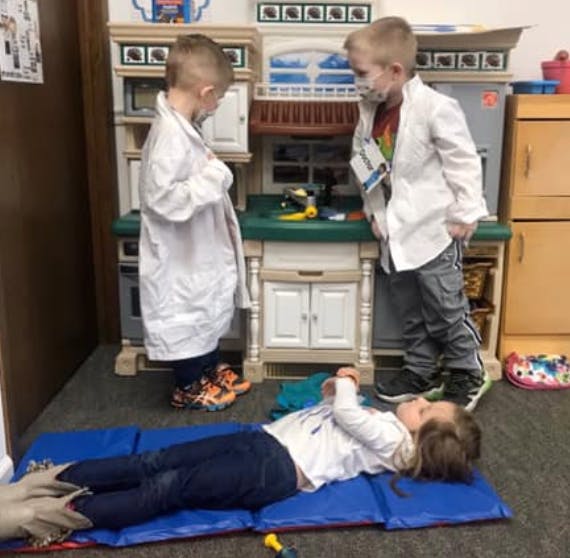 Two children in lab coats looking at a child lying on a mat