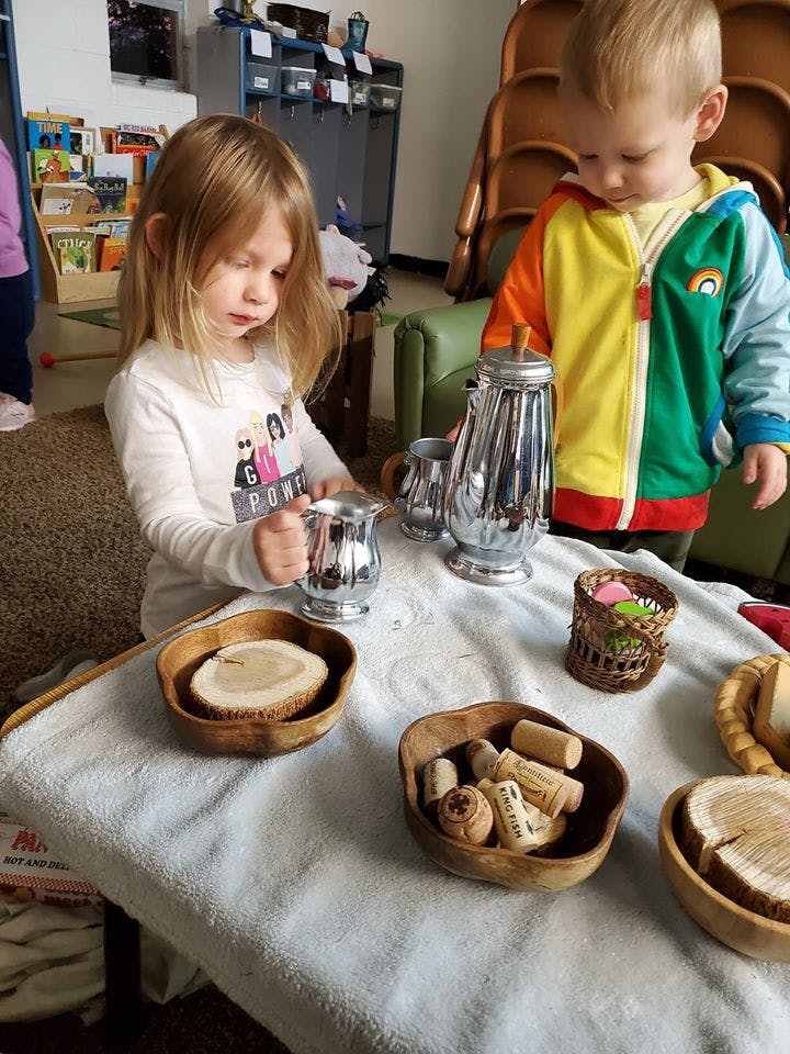 Child holding a silver pitcher at a table with another child nearby