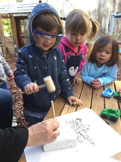 Child in safety glasses using a mallet to chip away at a block, with other children watching