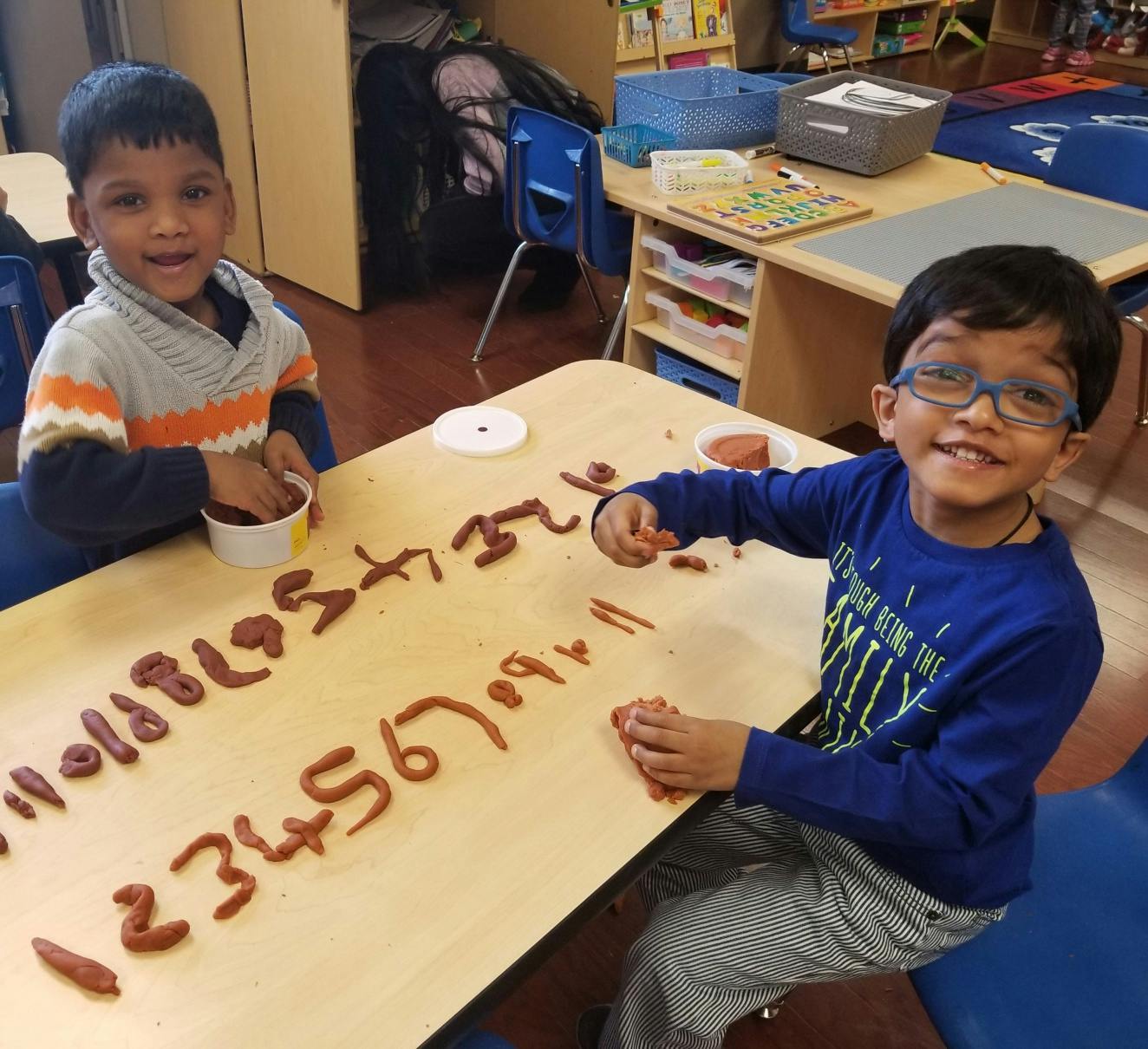 Two children playing with clay on a table, forming numbers and letters