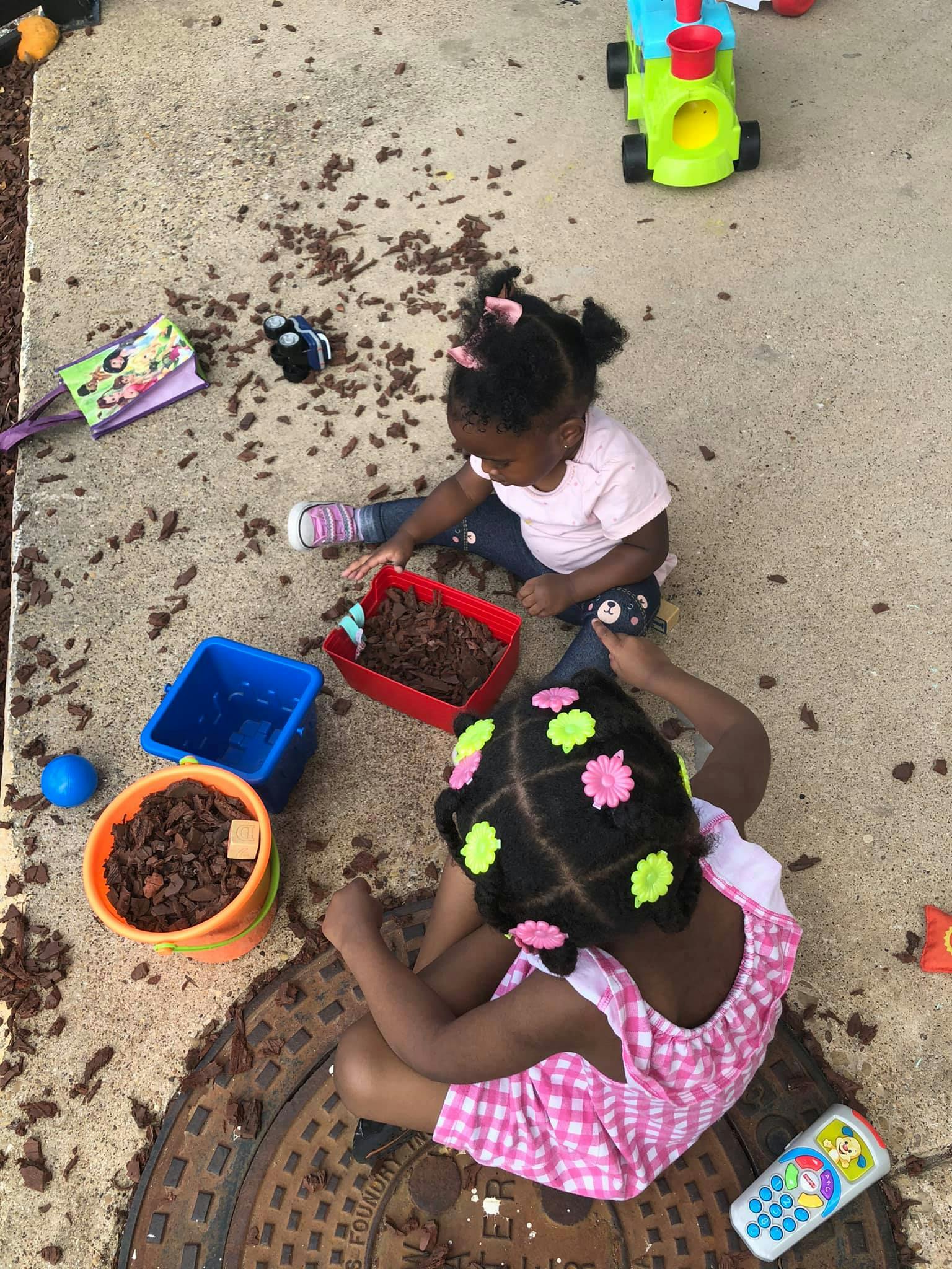 Two children playing with wood chips and toys on the ground