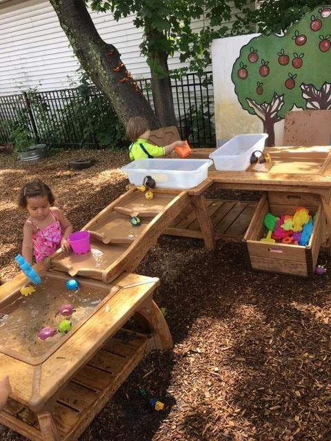 Two children playing with water and toys at a wooden activity table outdoors