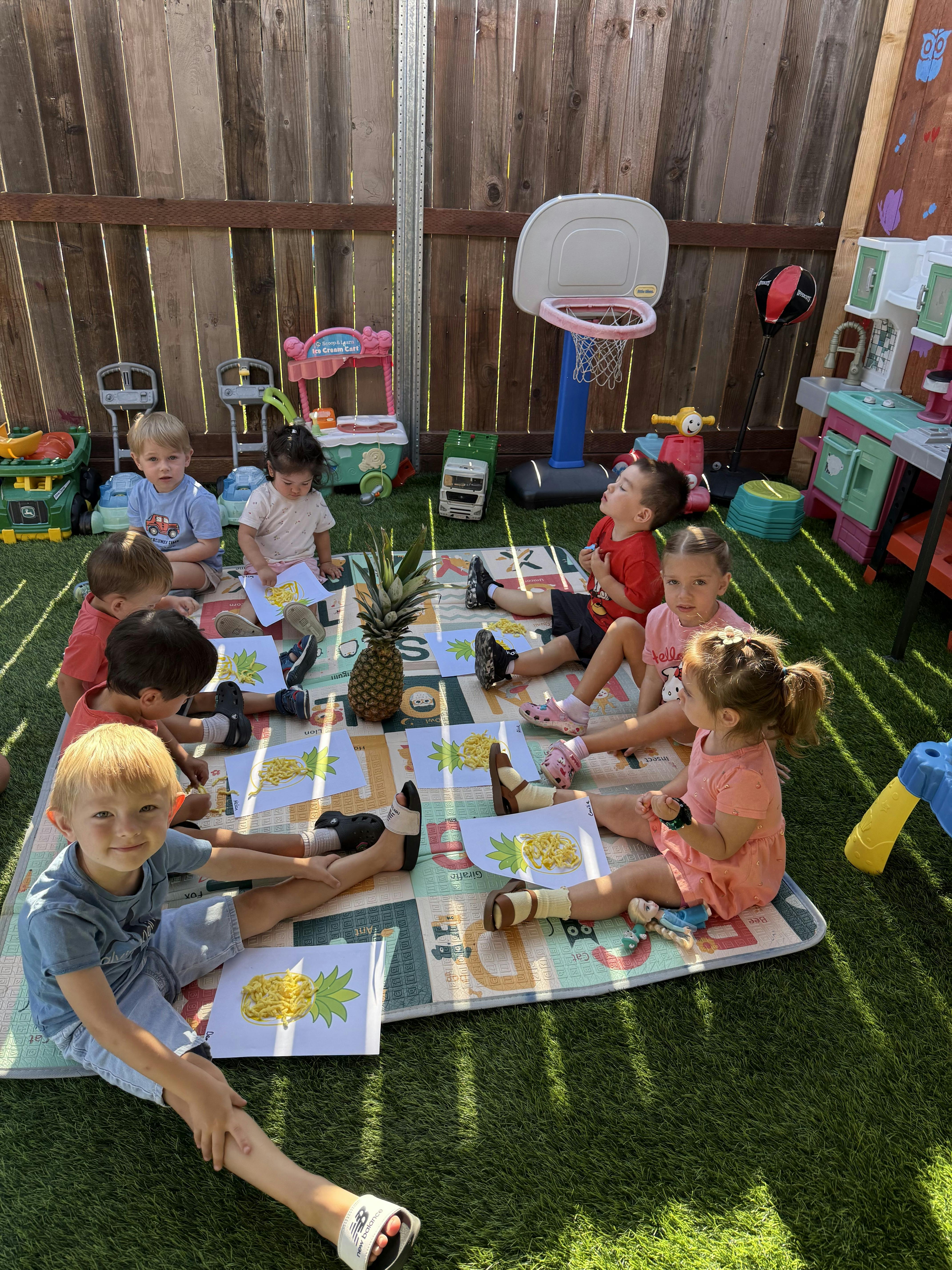 Group of children sitting on a mat outdoors crafting with pineapple illustrations