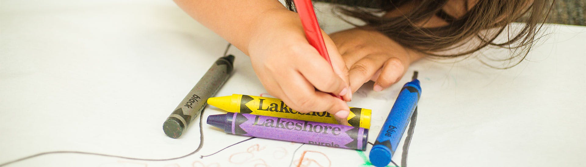 Child's hand drawing with a red crayon next to other crayons