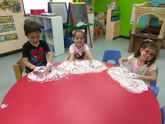 Three children playing with shaving cream on a red table in a classroom