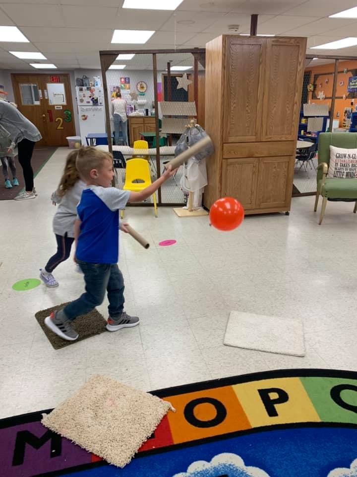 Child swinging a cardboard tube at an orange balloon in a classroom