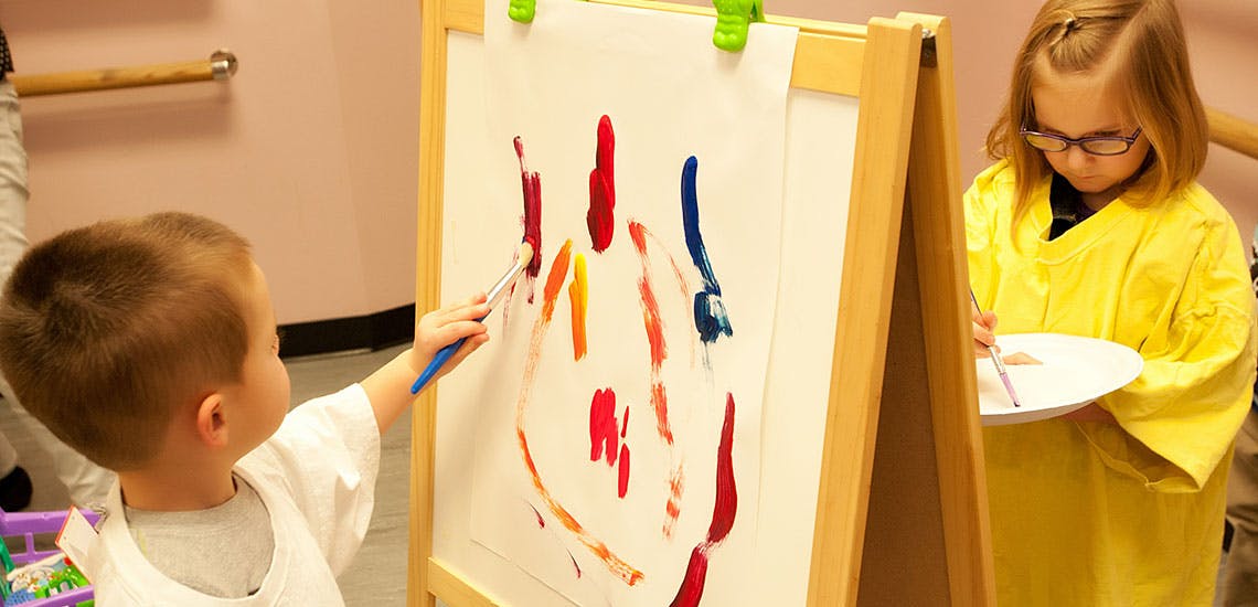 Child painting on an easel next to another child holding a palette