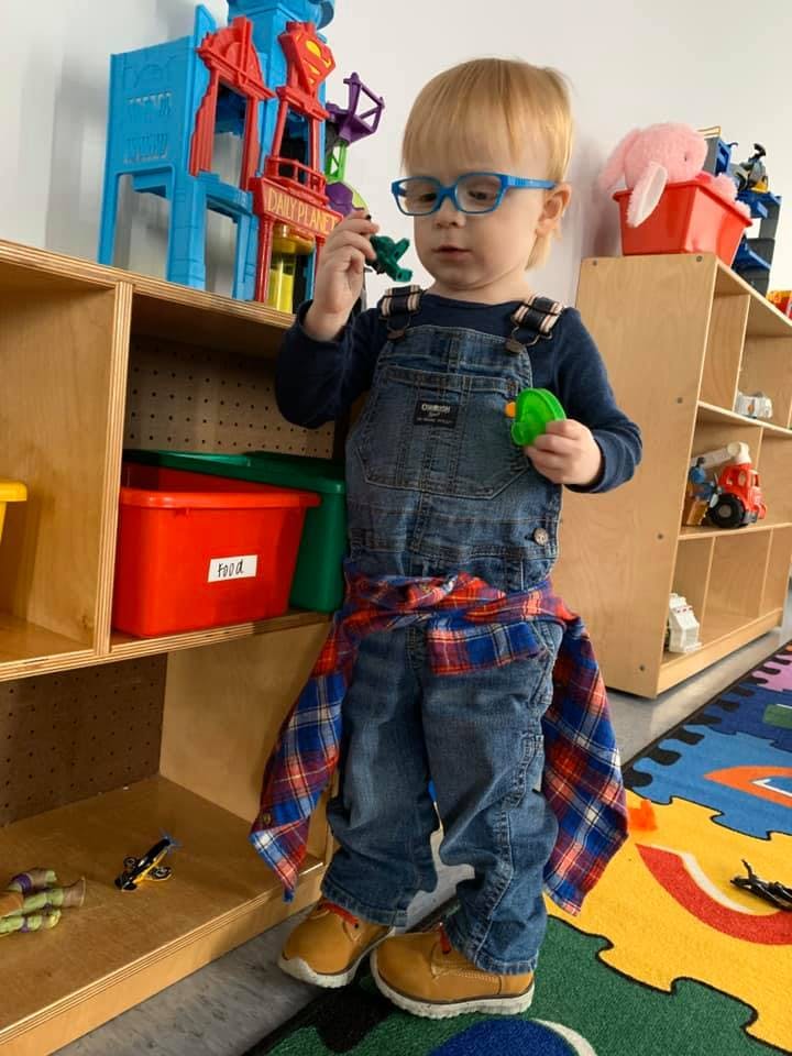 Child in overalls and glasses holding small toys in a classroom