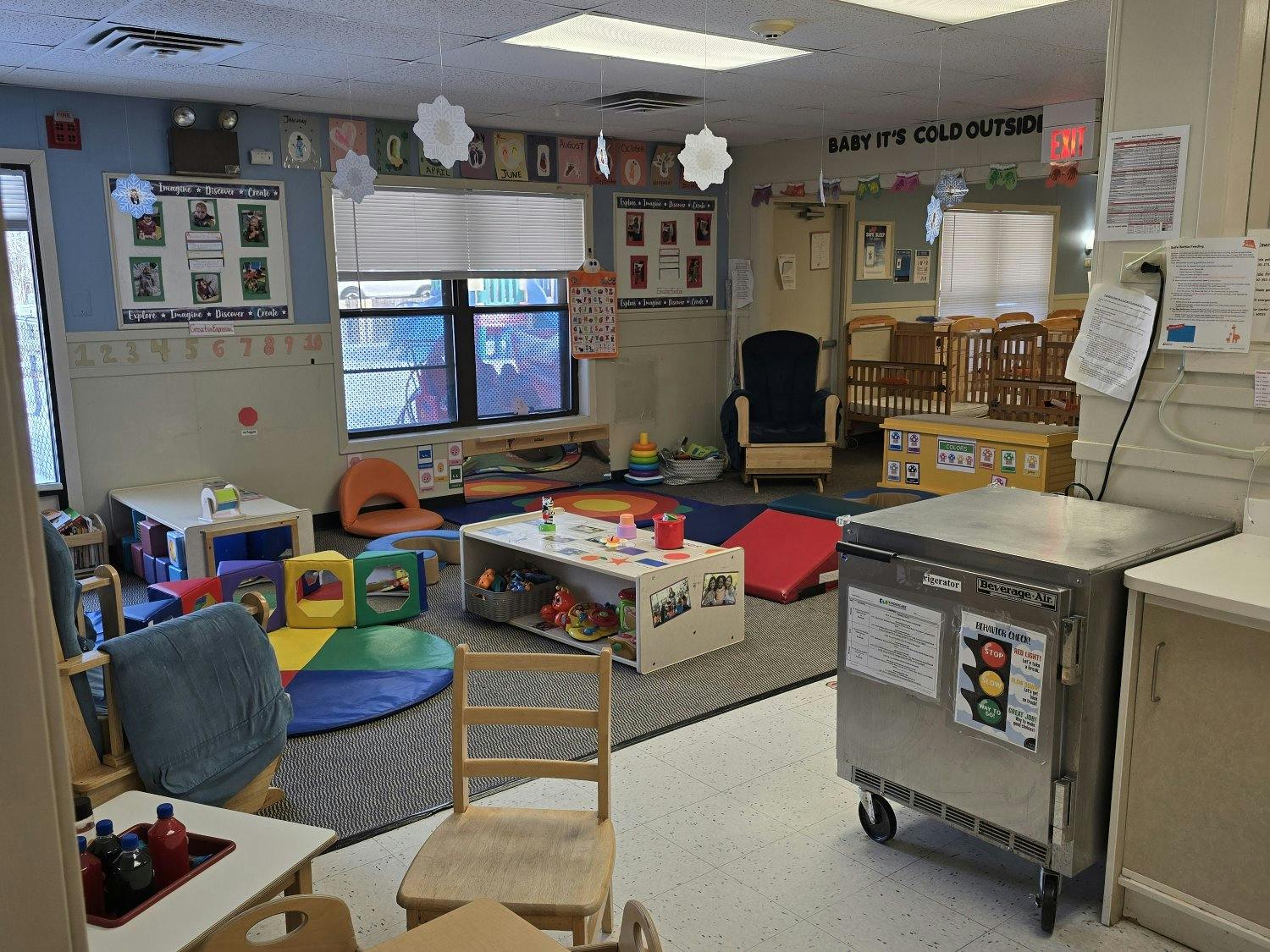 An empty classroom with various play areas, cribs, and a beverage refrigerator