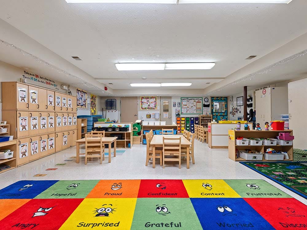 Empty classroom with small tables and chairs, cubbies, and a colorful rug with emotions