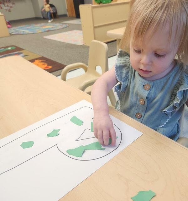 Child placing green paper shapes on a paper outline on a table