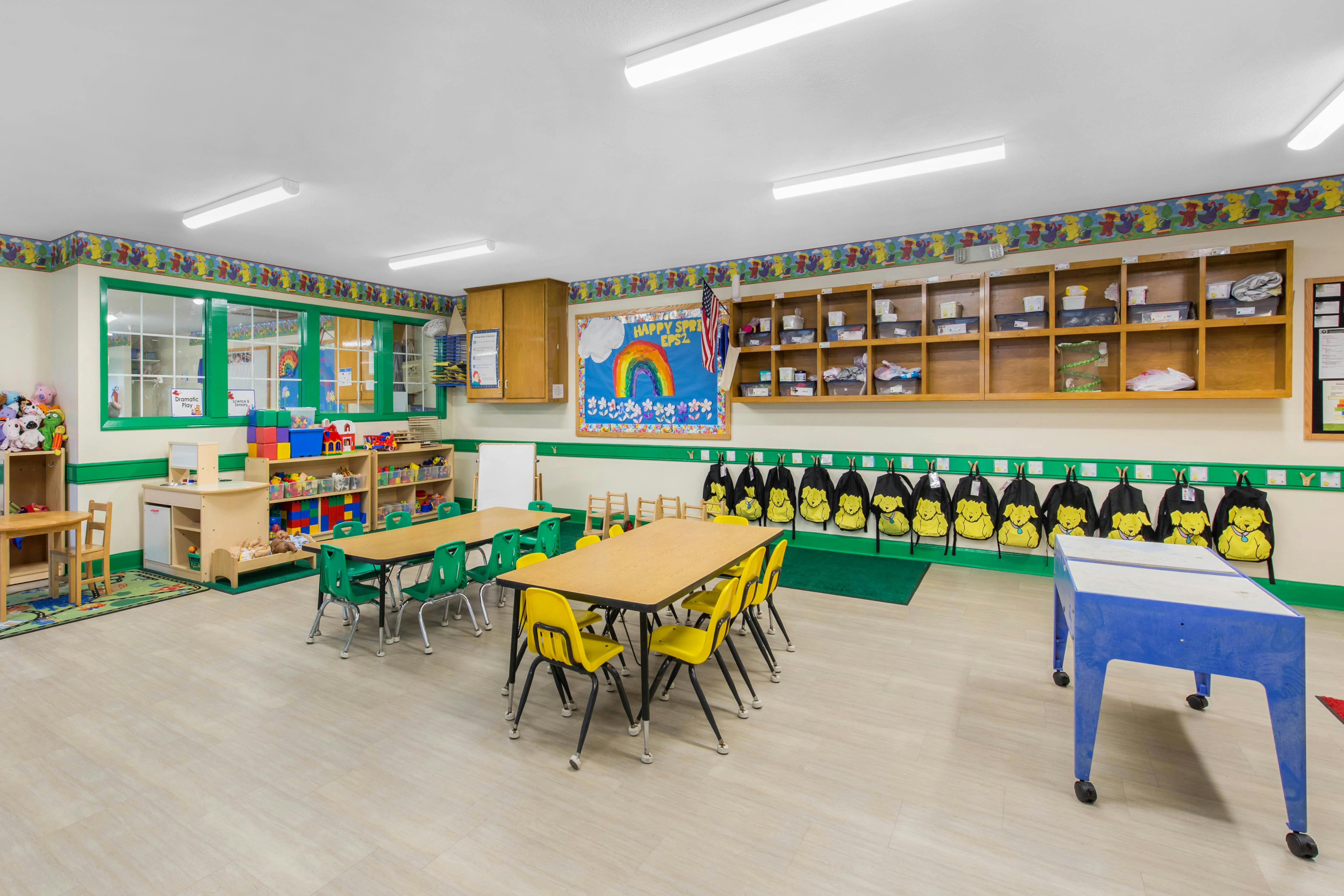 Classroom with child-sized tables and chairs, shelves with toys, and backpacks