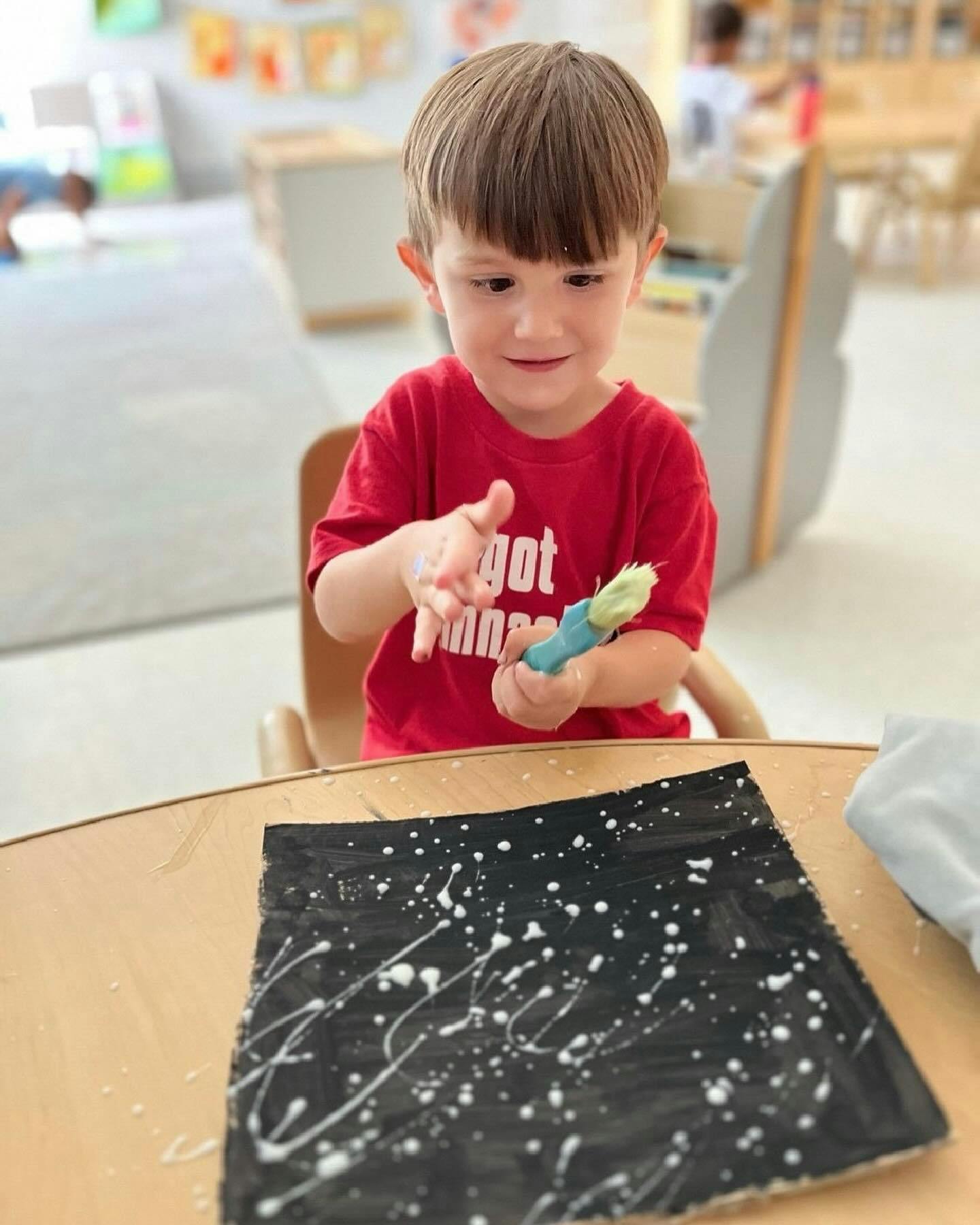 Child holding a brush over a black paper with white paint splatters