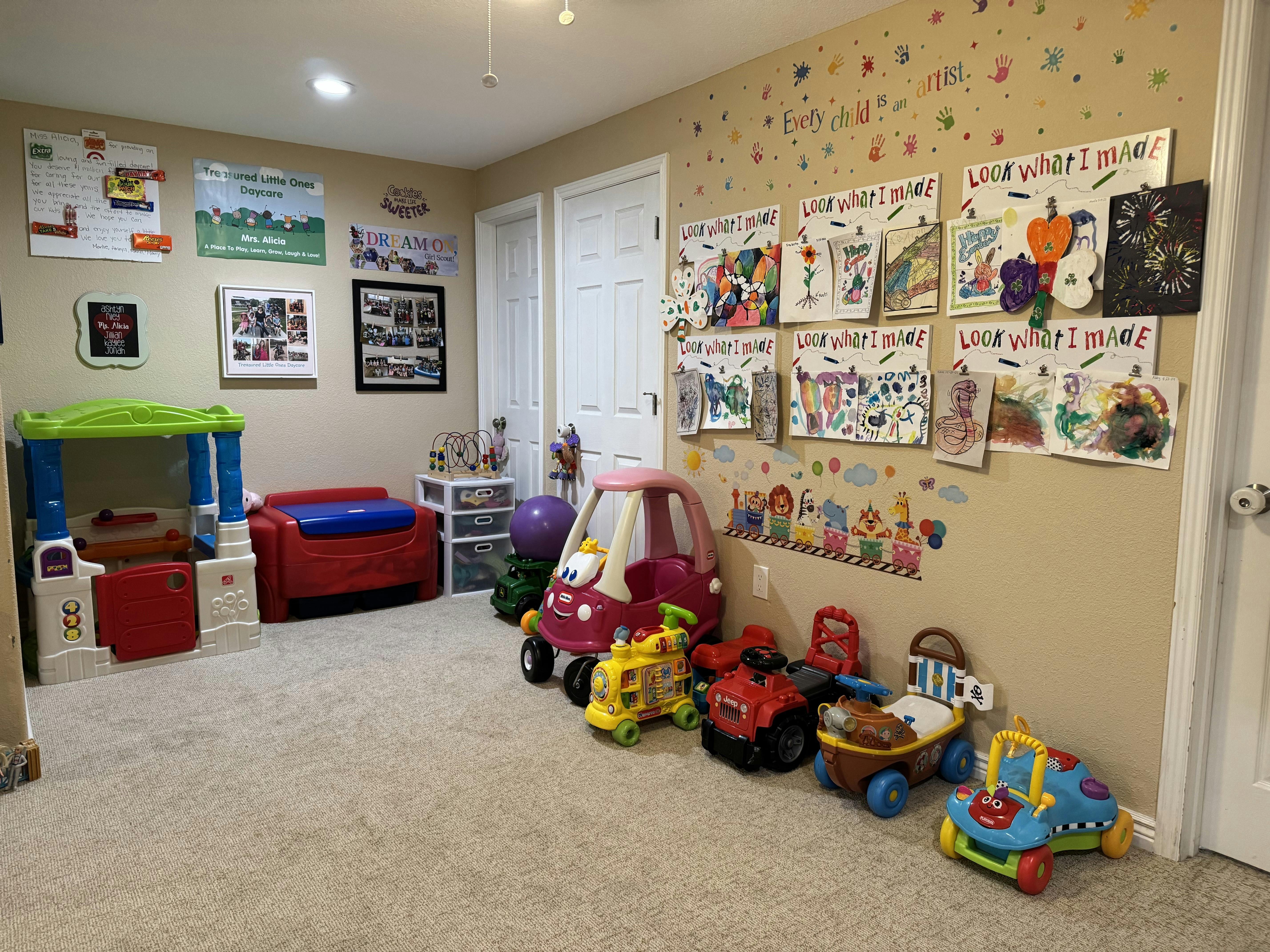 A playroom with various ride-on toys, children's artwork displayed on the wall, and signs for a daycare