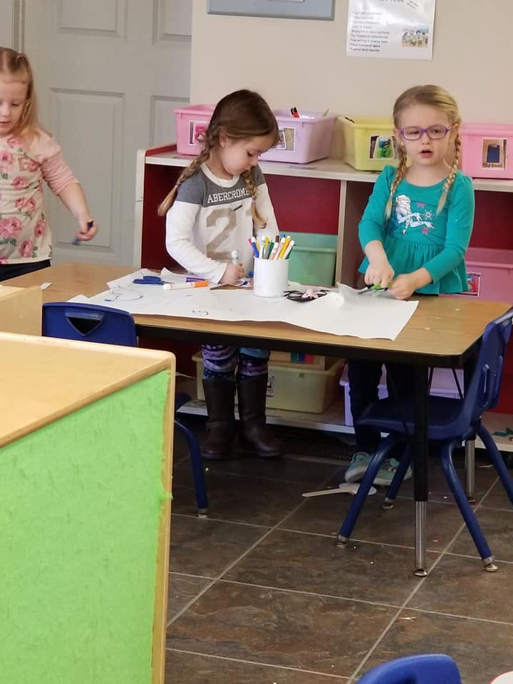 Three children in a classroom, two at a table with art supplies