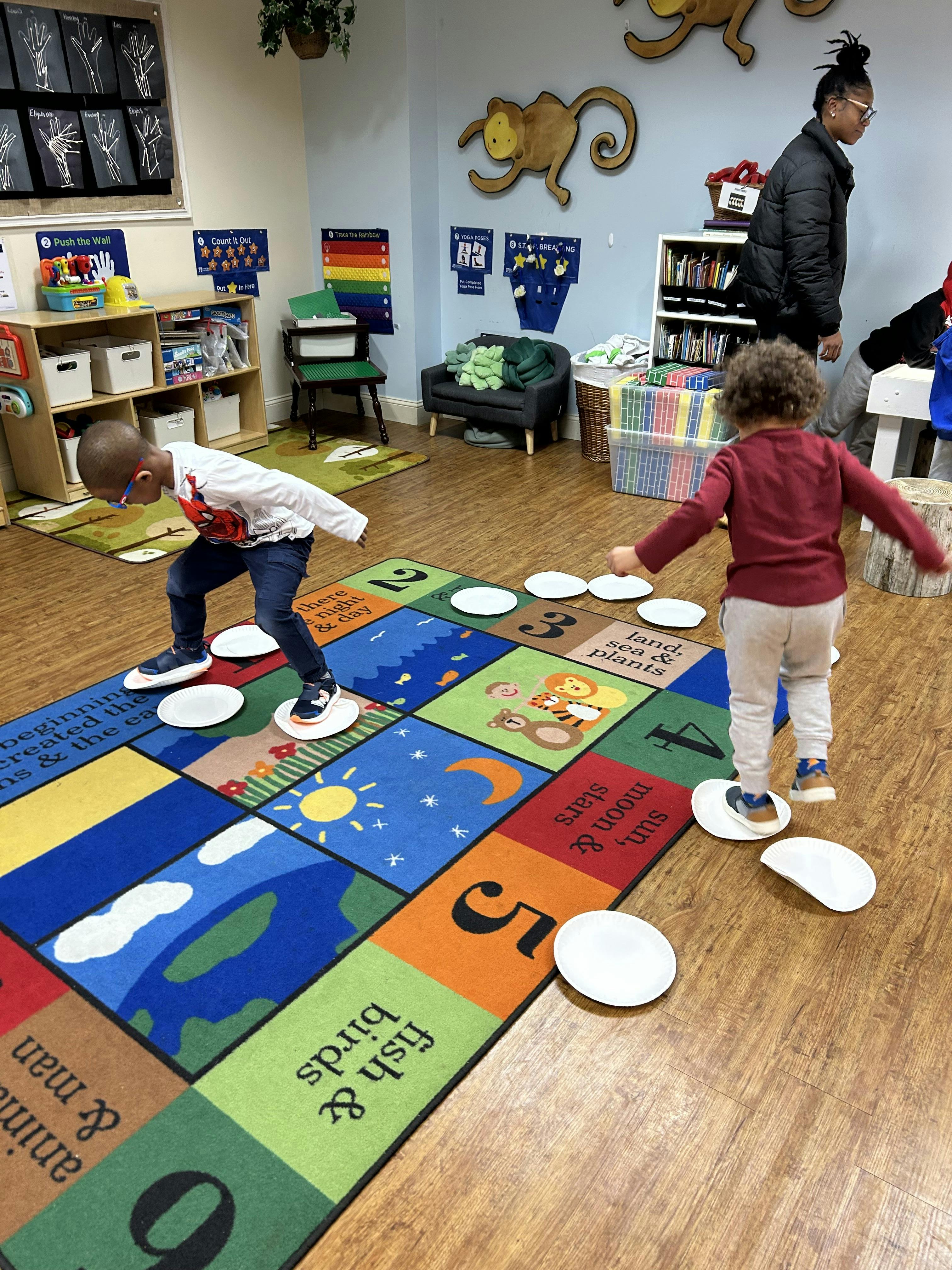 Two children stepping on paper plates on a patterned rug in a classroom