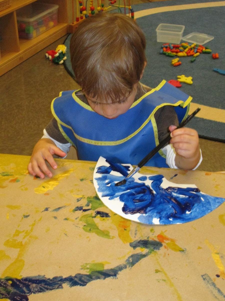 Child in a blue smock painting a paper plate with blue paint