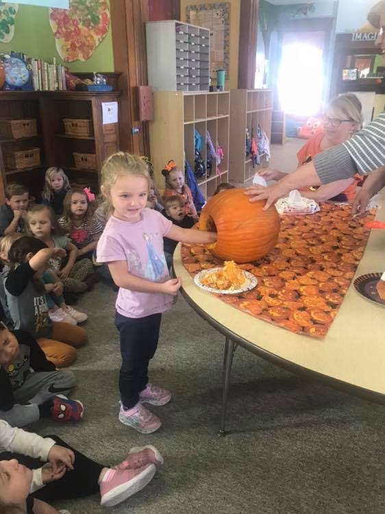 Child standing next to a table with a pumpkin, other children sitting on floor