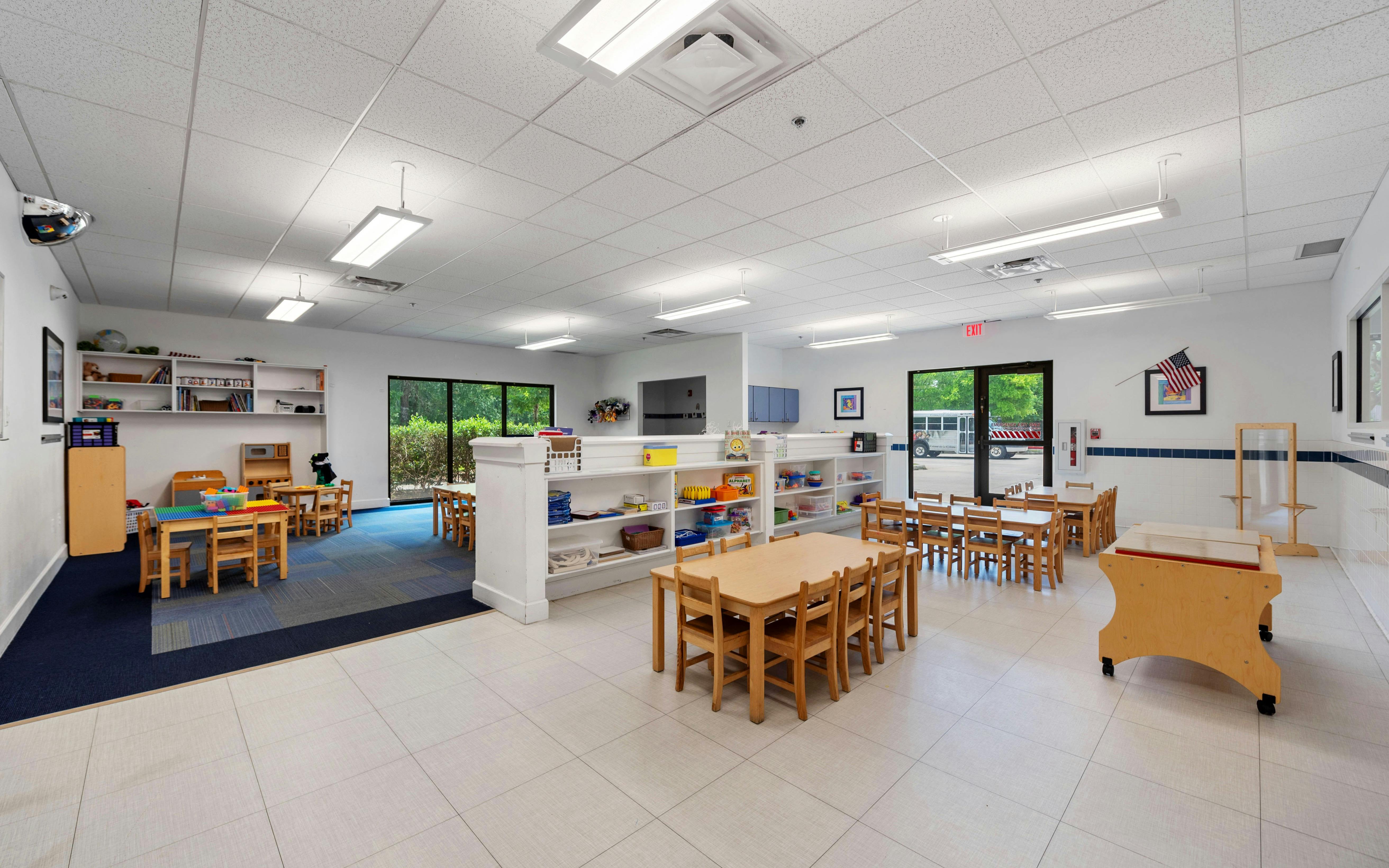 Empty classroom with small wooden tables and chairs, shelving with toys and books