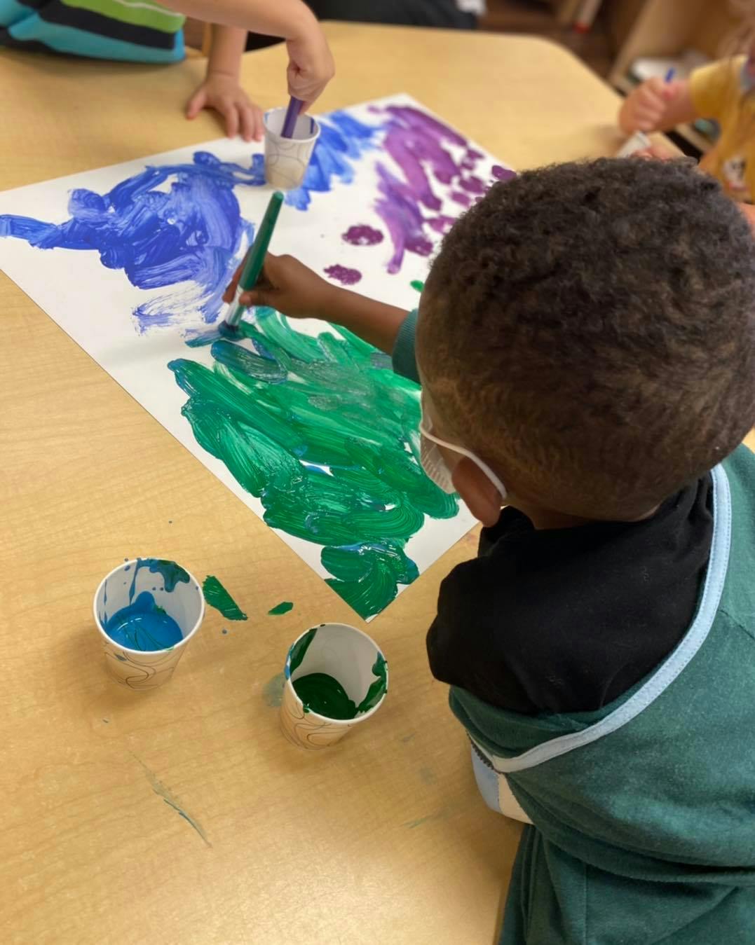 Child painting on paper with green, blue, and purple paint