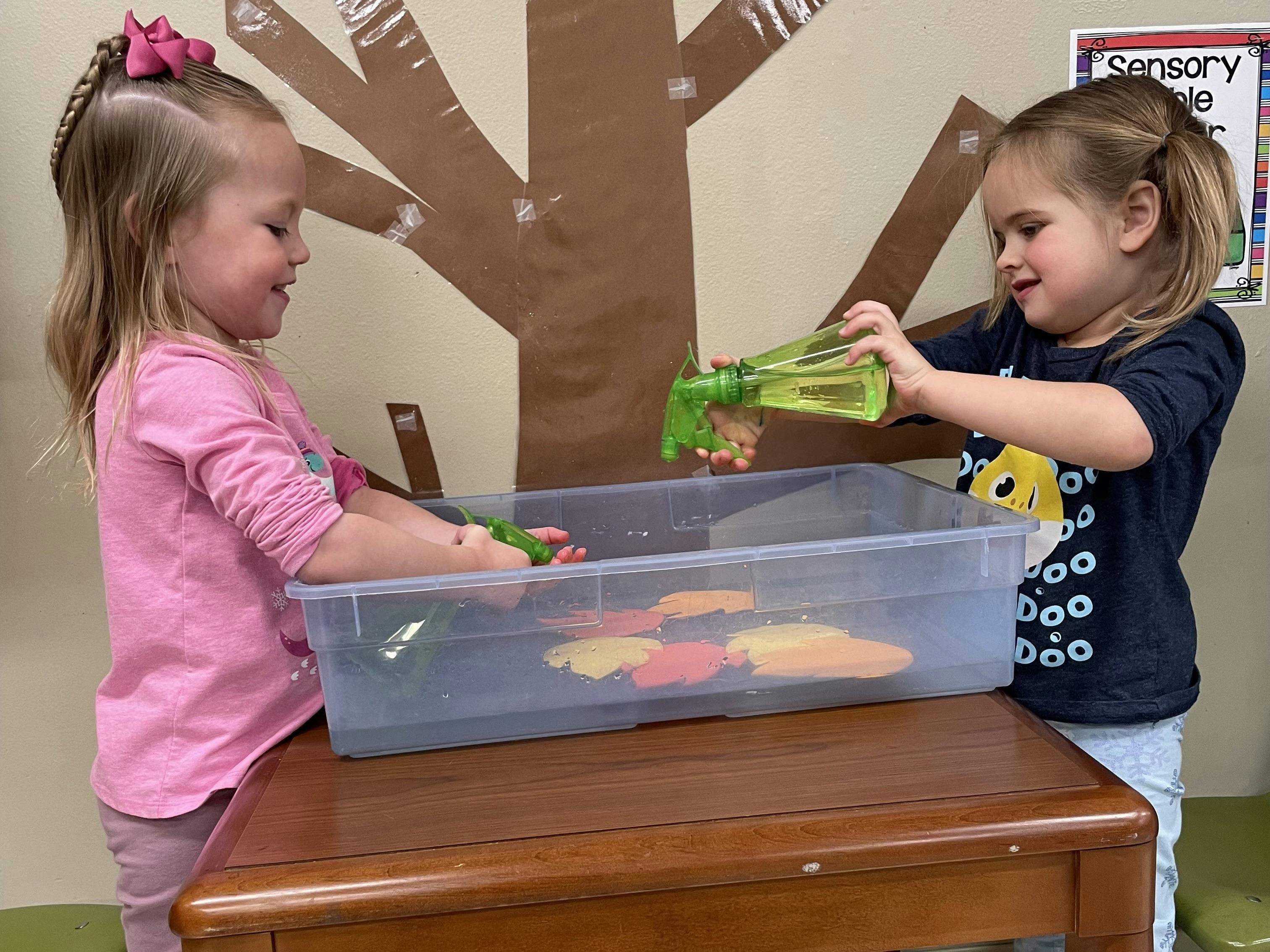 Two children spraying water into a bin with shapes
