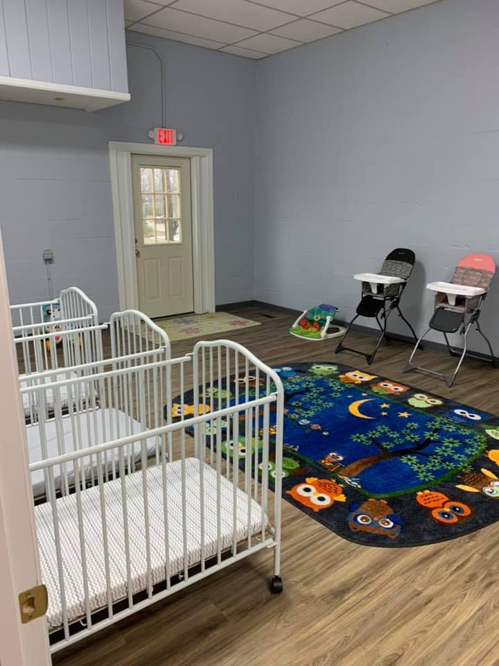 Three white cribs in a room with a colorful rug and two high chairs