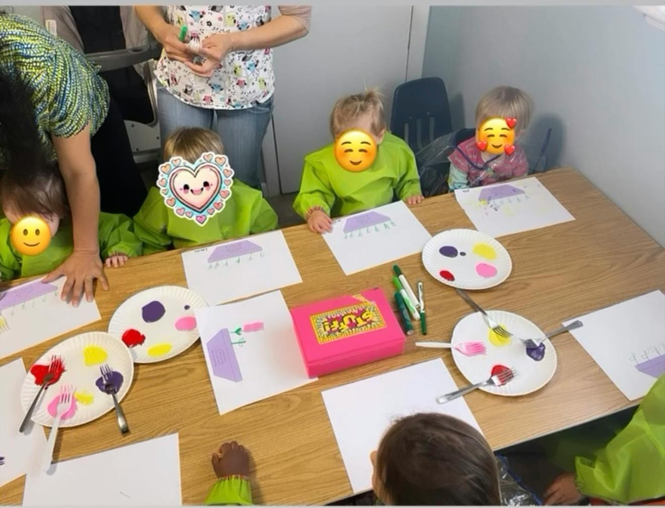 Children in green smocks painting at a table with paper plates of paint and forks