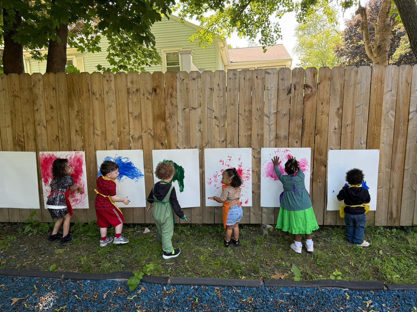 Children finger painting on papers attached to an outdoor wooden fence