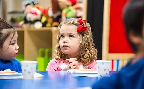 Child with curly hair and red bows looking up at a table with other children