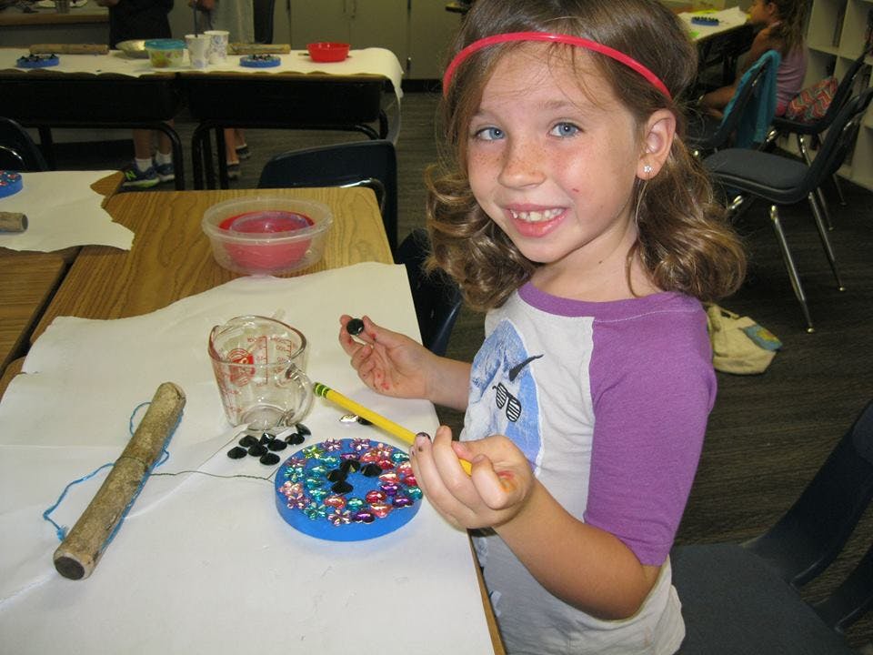 Child holding a bead and a yellow pencil, with craft supplies on a table