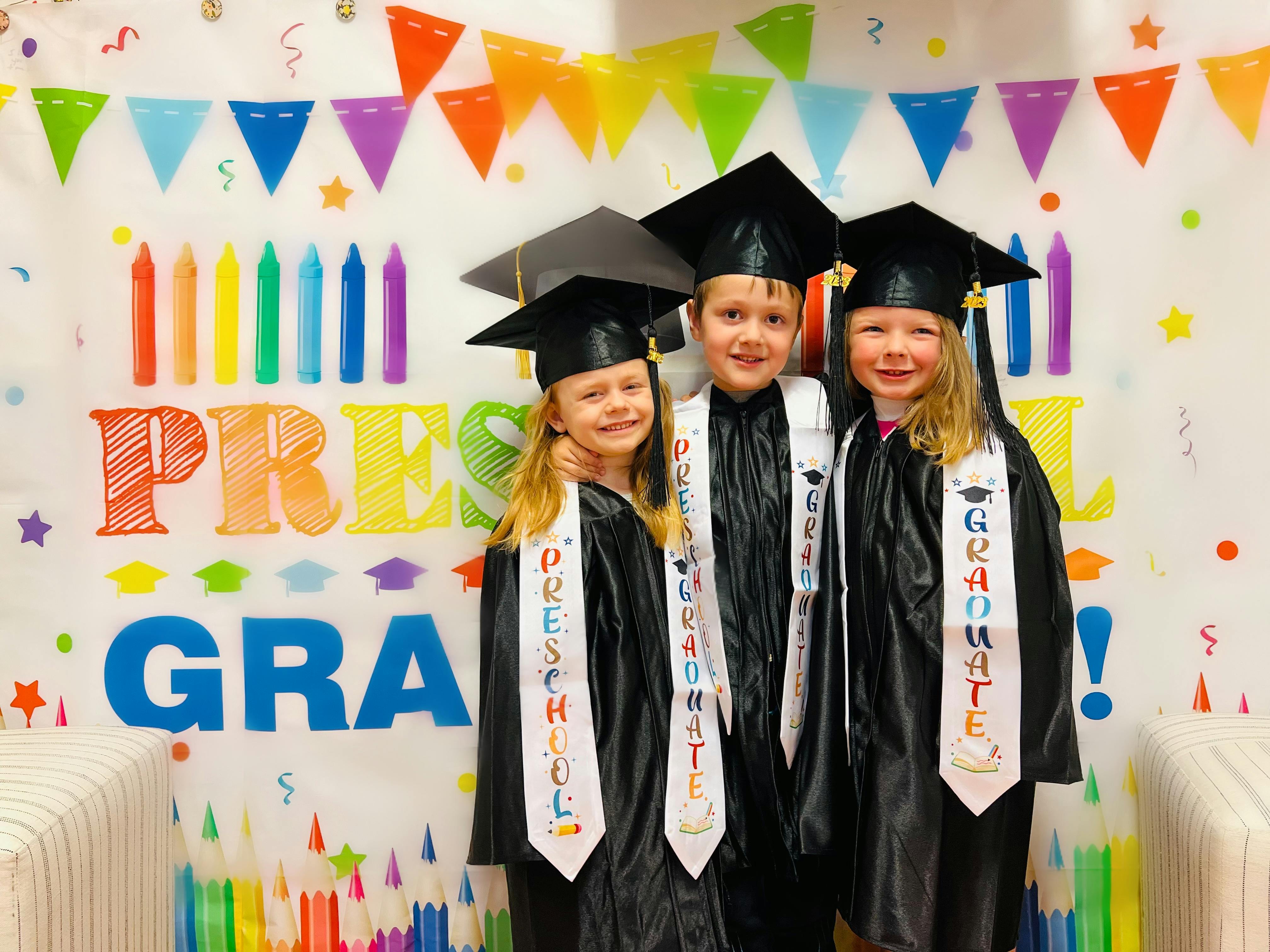 Three children in graduation caps and gowns with sashes that say "Preschool Graduate"