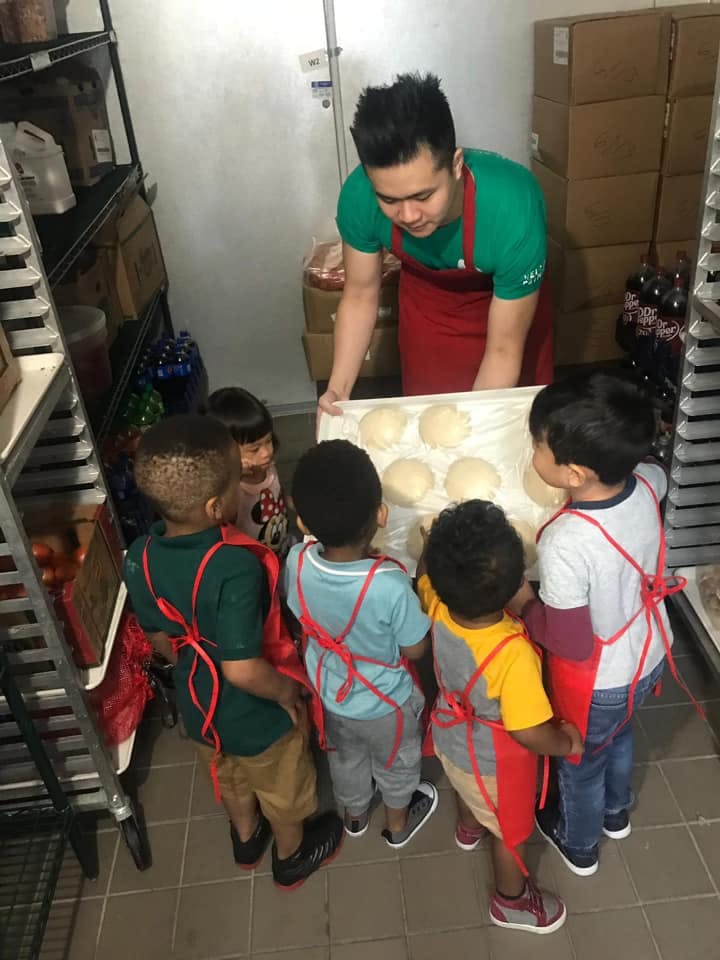 Person in apron showing children a tray of dough