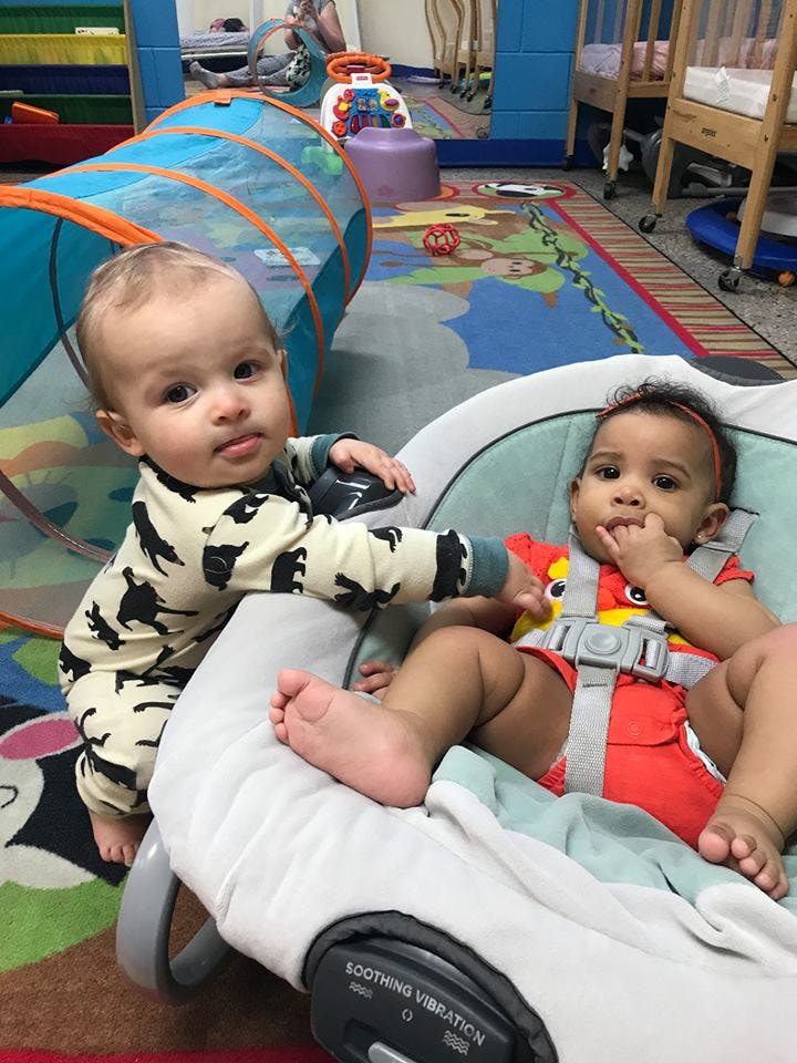 Two children playing in a room, one standing and one sitting in a bouncer