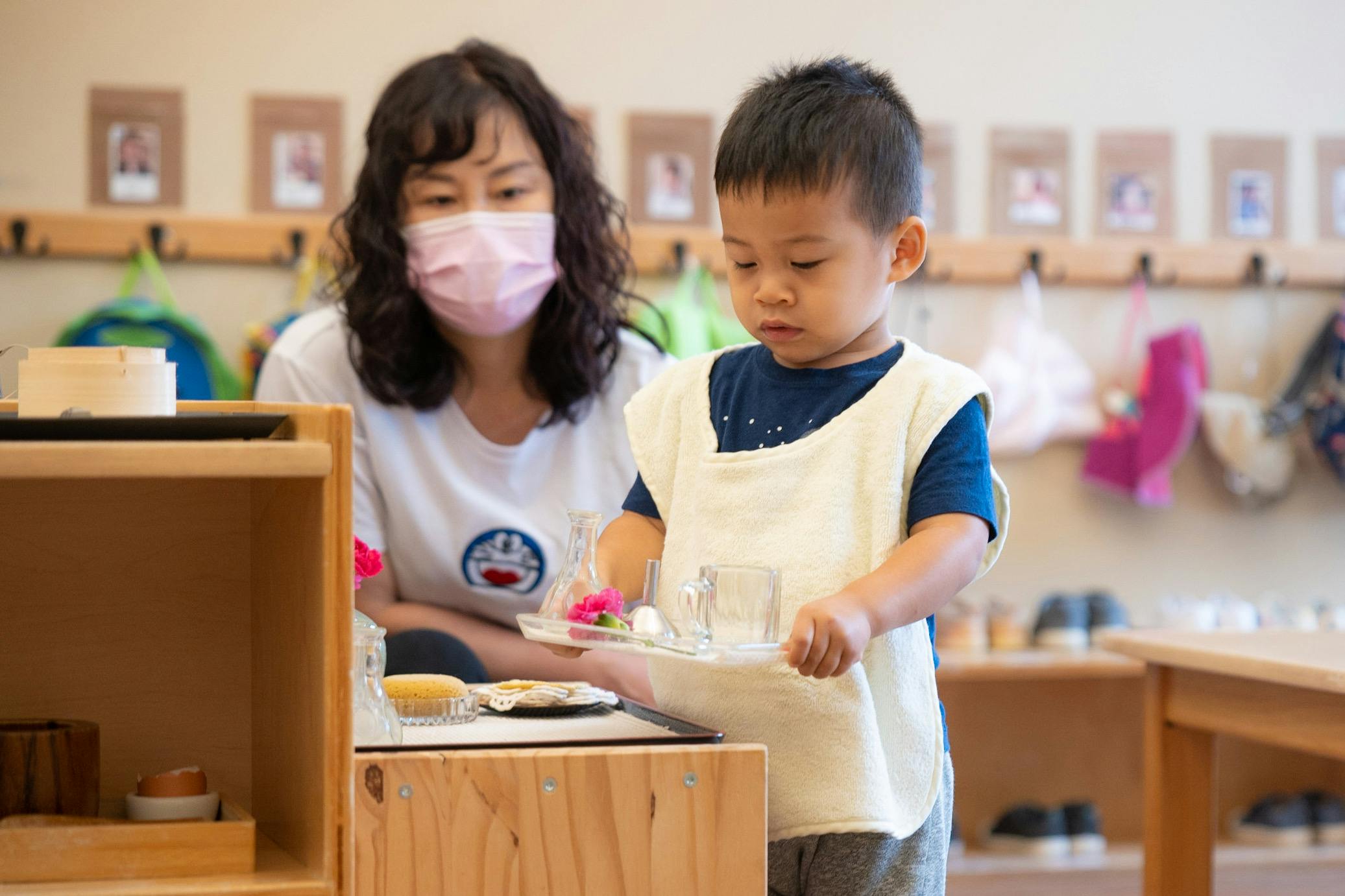 Adult in mask and child standing at a wooden counter in a classroom