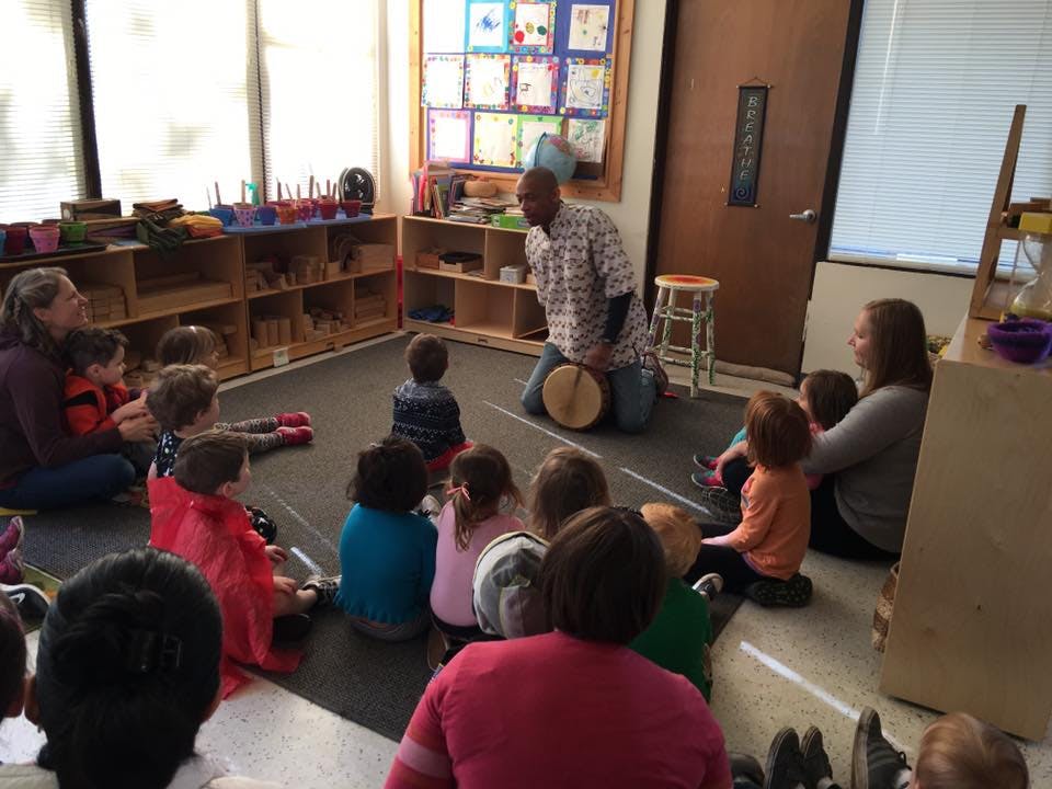 Person kneeling with a drum in front of children and adults in a classroom