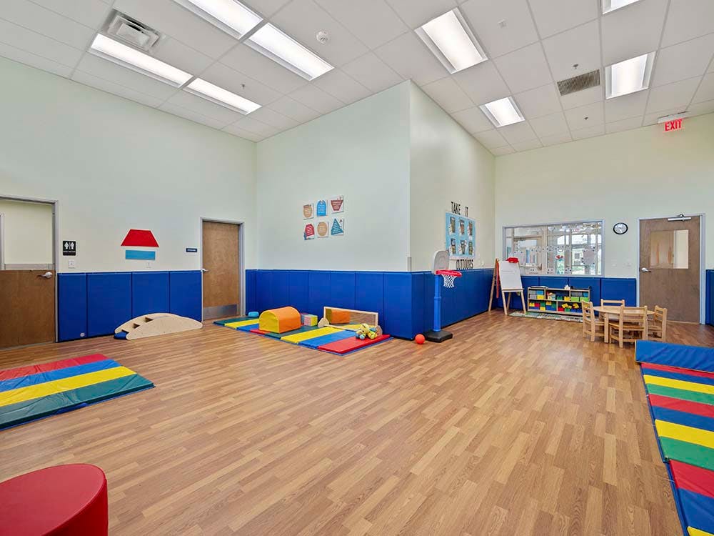 An empty classroom with colorful mats, a small table and chairs, and a basketball hoop