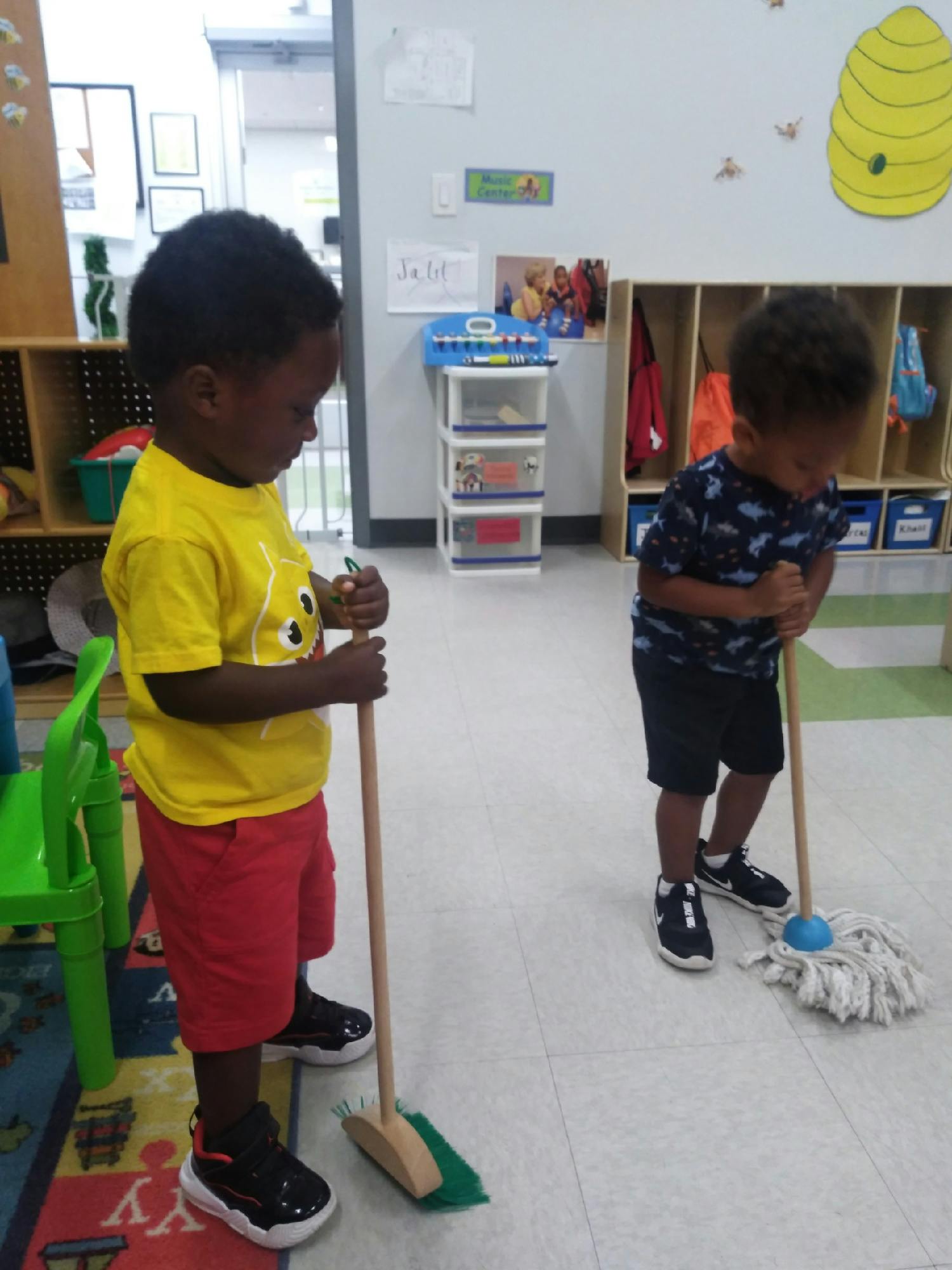 Two children holding a toy broom and mop in a classroom
