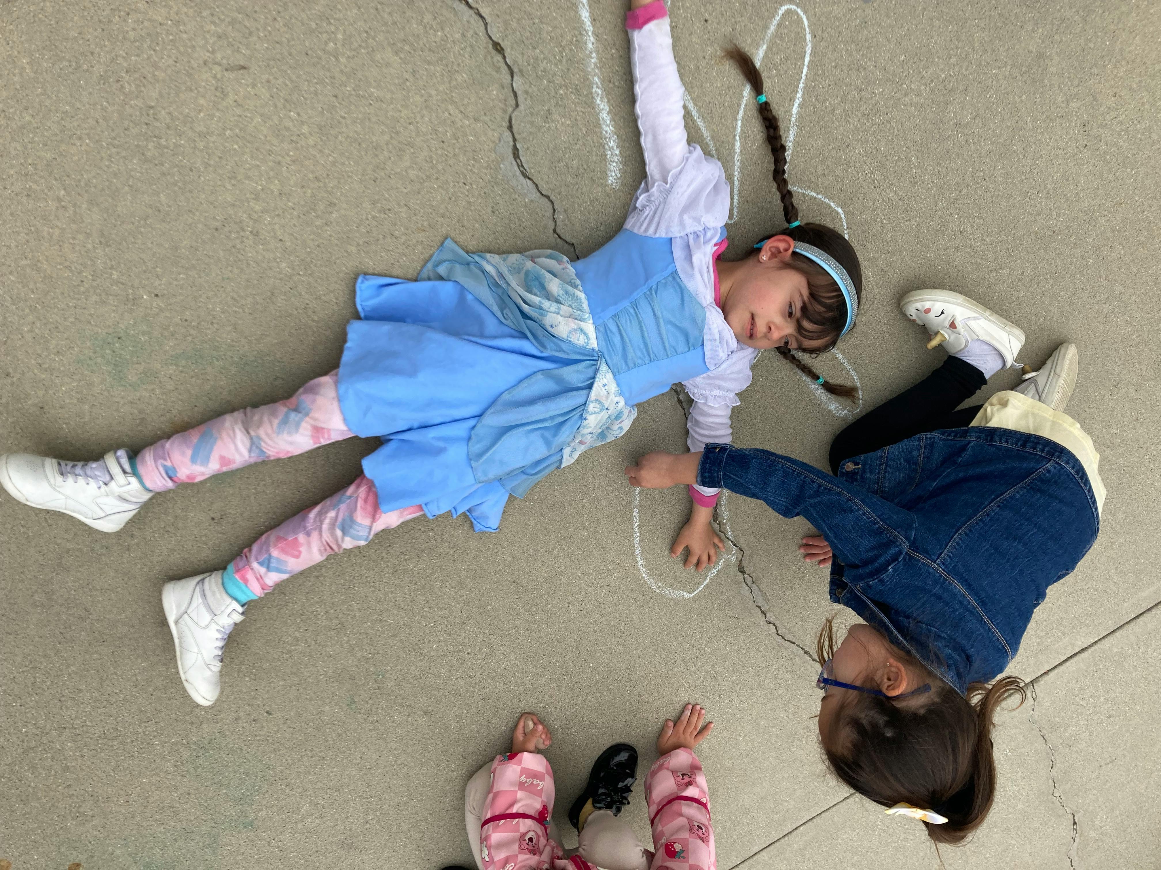 Child in blue dress lying on ground with chalk outline, another child nearby