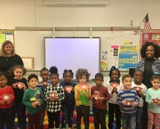 Two adults with a group of children holding up crafts in a classroom
