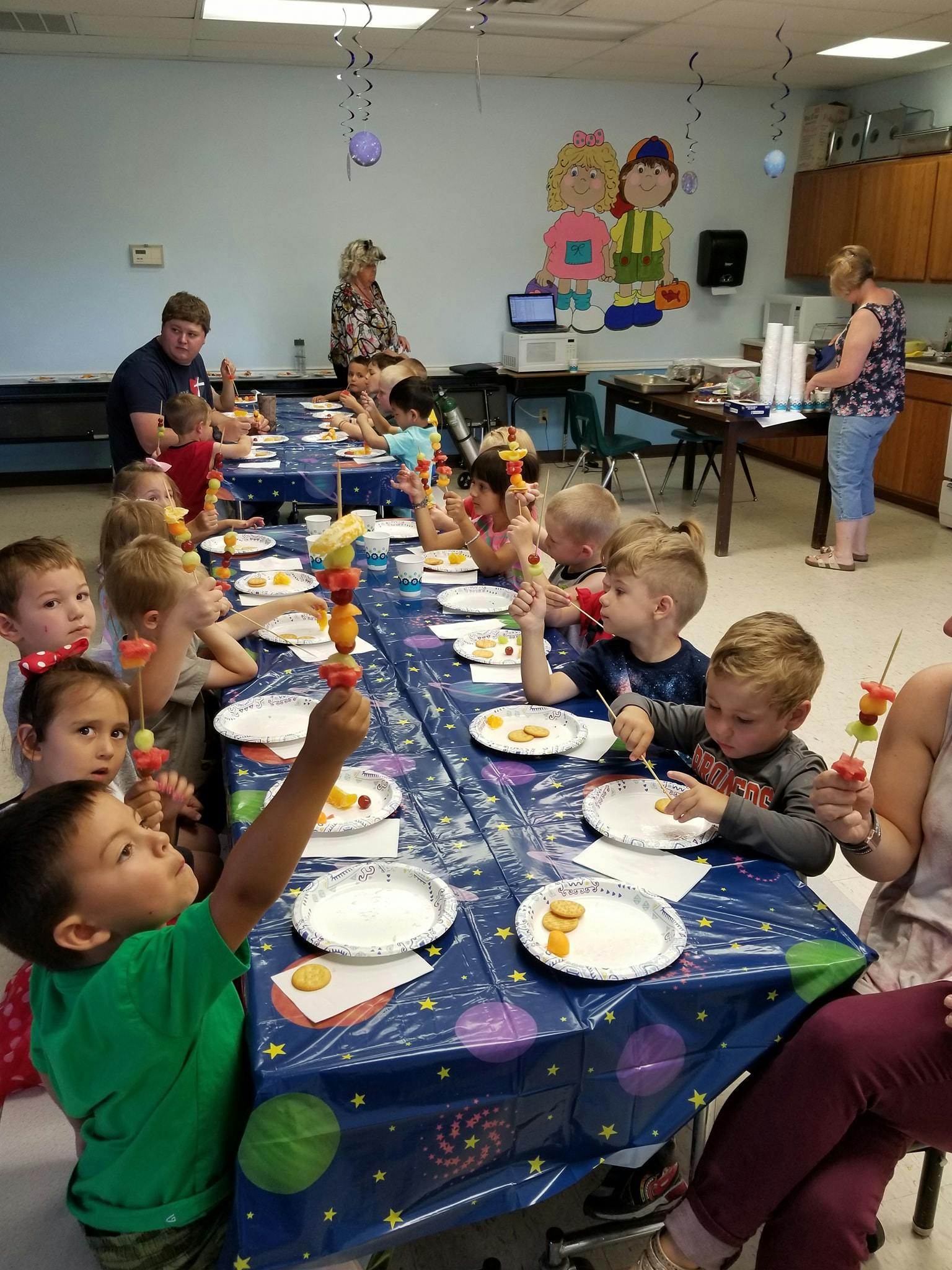 Children and adults sitting at a long table with fruit skewers