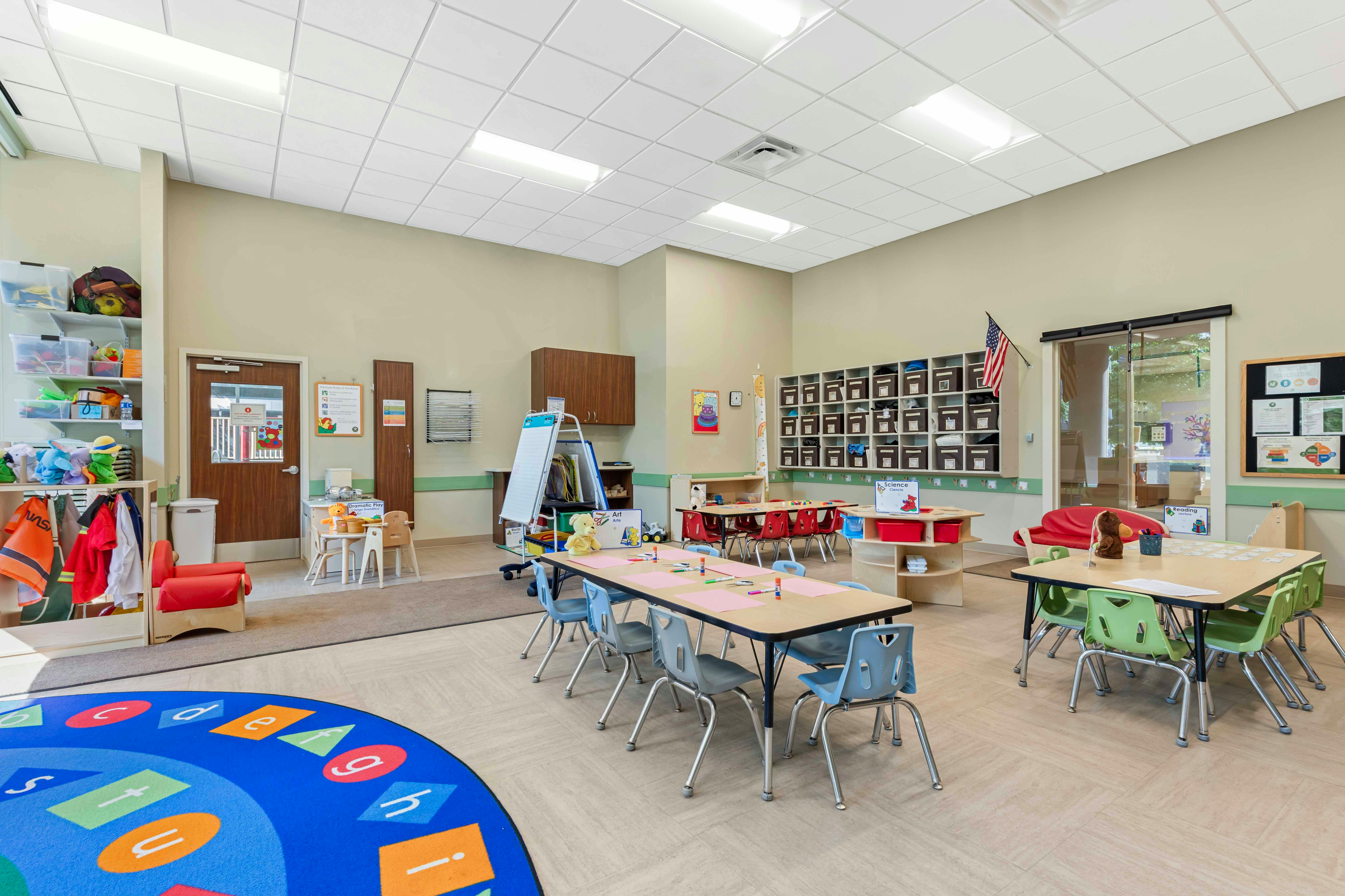Classroom with multiple tables and chairs, shelves, and a colorful rug