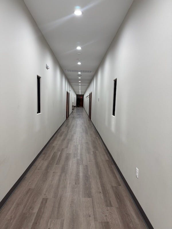 A long, narrow hallway with light-colored walls, recessed lighting, and wood-look flooring