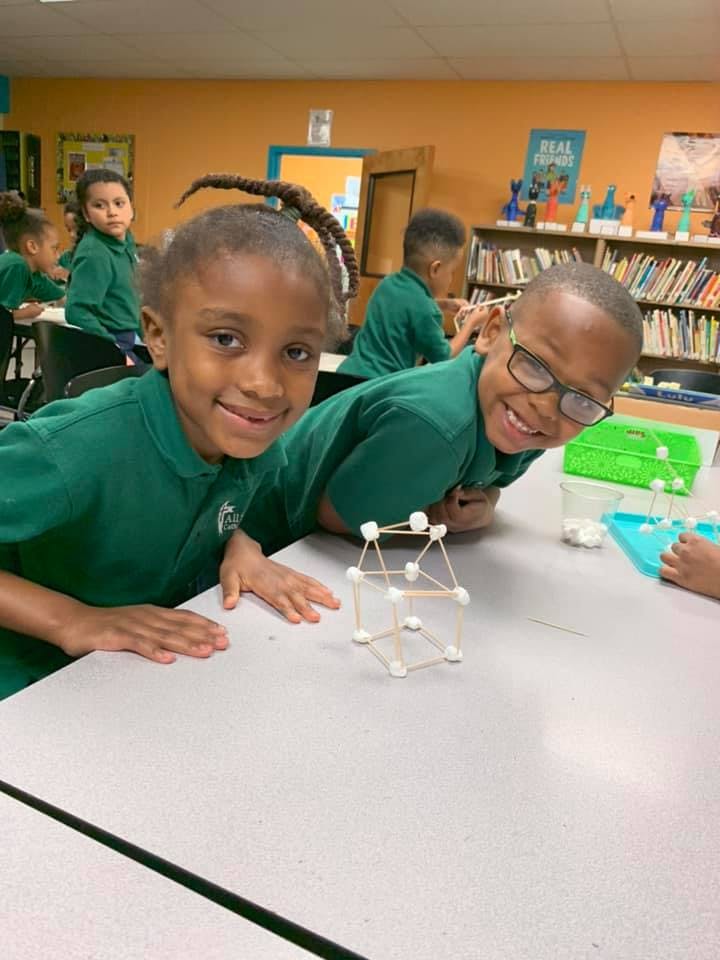Two children in green shirts building with marshmallows and toothpicks in a classroom