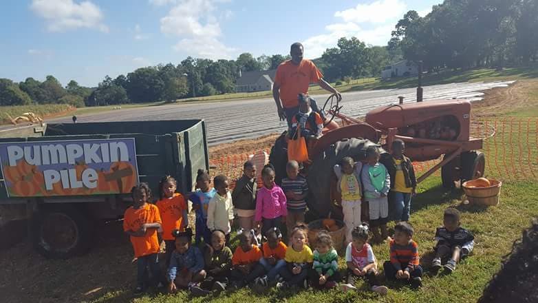 Person on a tractor with children gathered around a trailer with a "Pumpkin Pile" sign