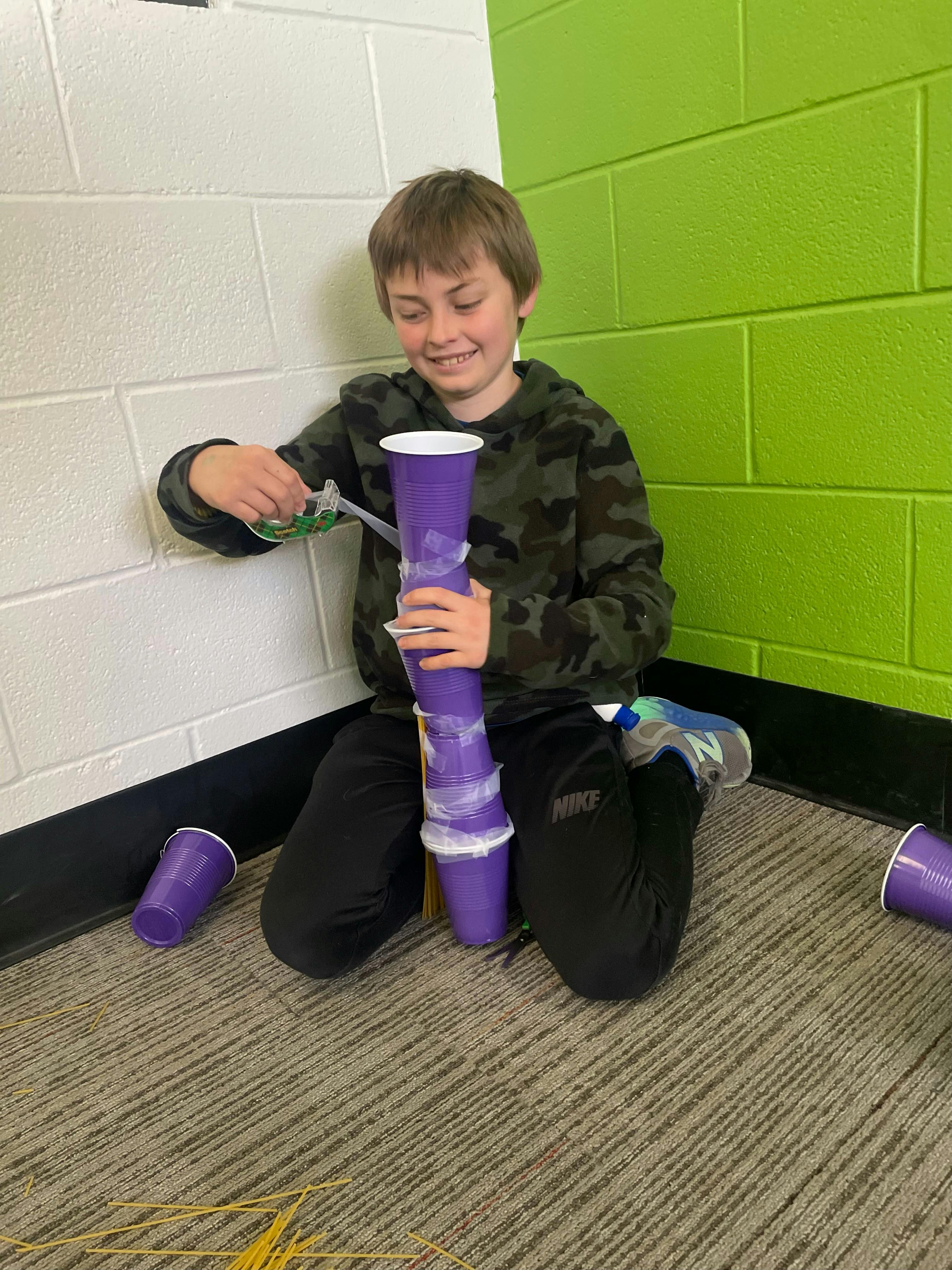 Child kneeling, building a tower of purple cups with tape