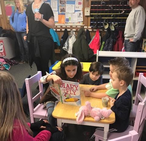 Child reading a book to other children at a table in a classroom
