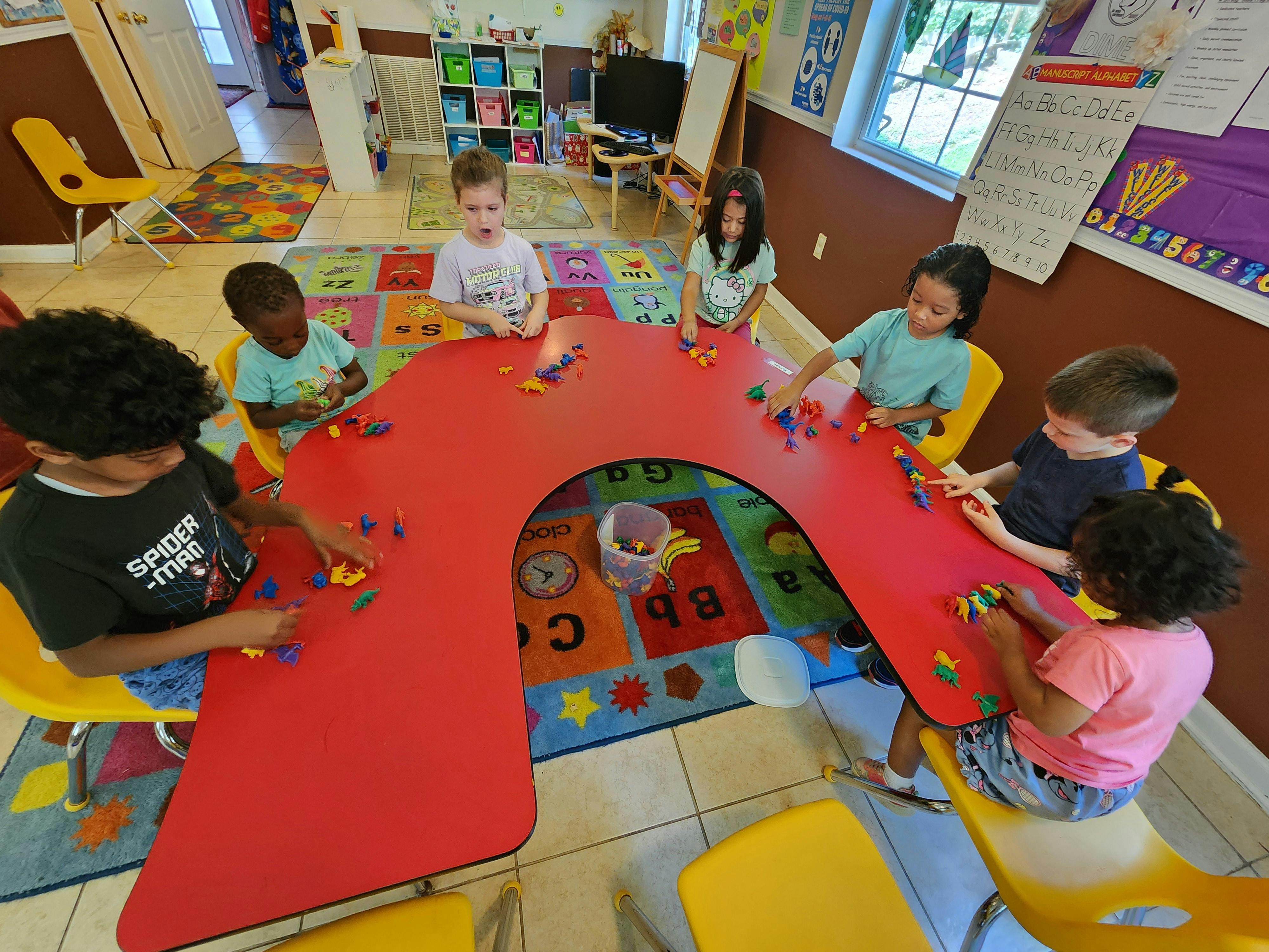 Group of children playing with animal toys at a red U-shaped table in a classroom